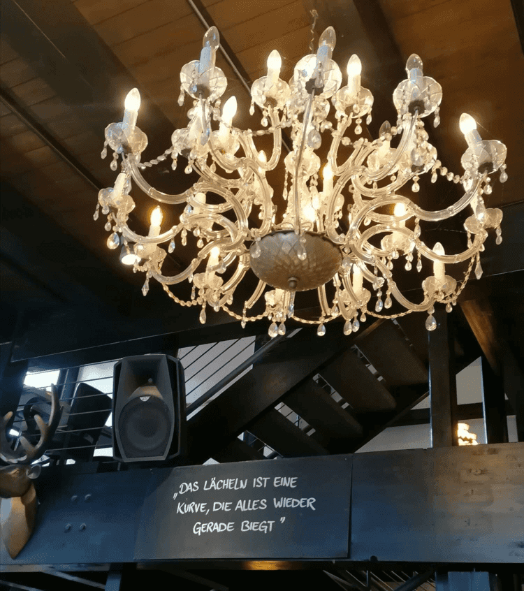 Ein Kronleuchter, darunter ist auf Holz ein Sinnspruch zu lesen. die schönste Kurve ist das Lächeln